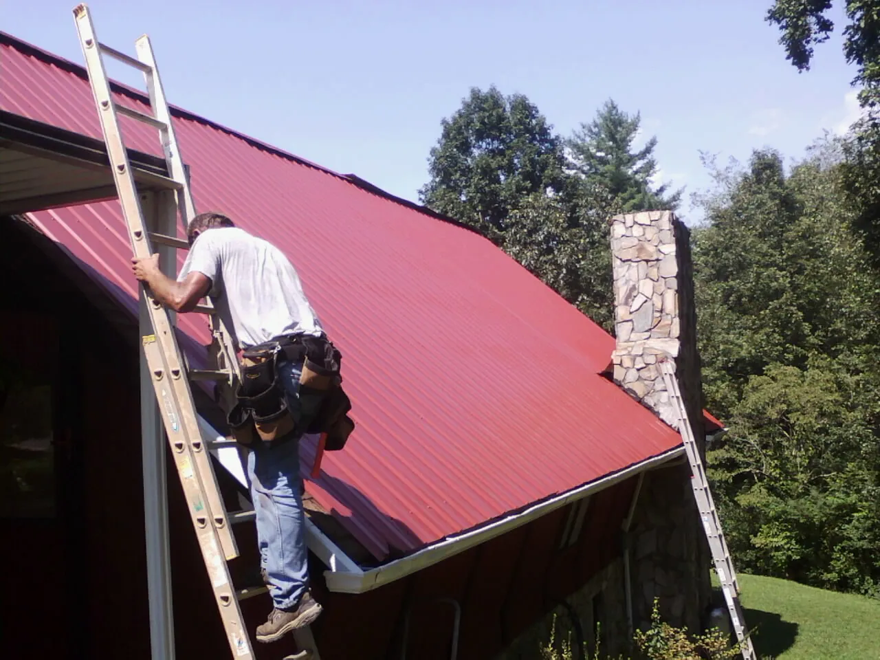Roofer on a ladder beside a red metal roof and stone chimney — Soffit & Fascia Repair by Roofing Brothers in Roosevelt